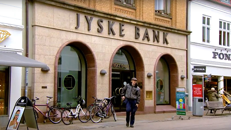 Person walks out of JYSKE Bank onto a road lined with bicycles, a bench, and store signs. 